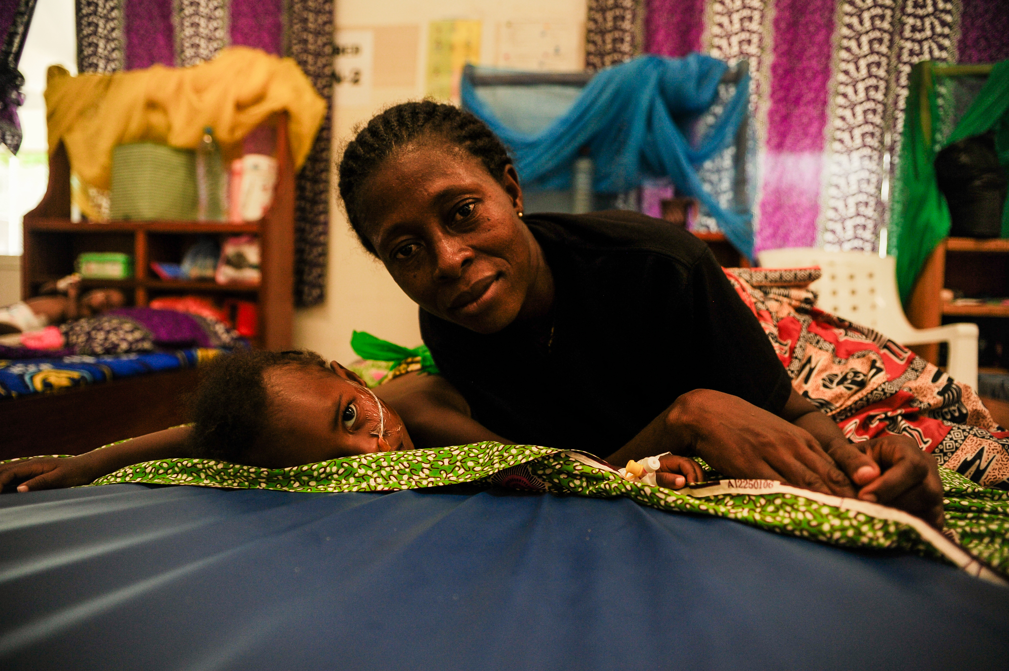 A caregiver smiles beside a child resting on an inpatient bed at the Jesus Loves Me Children's Center