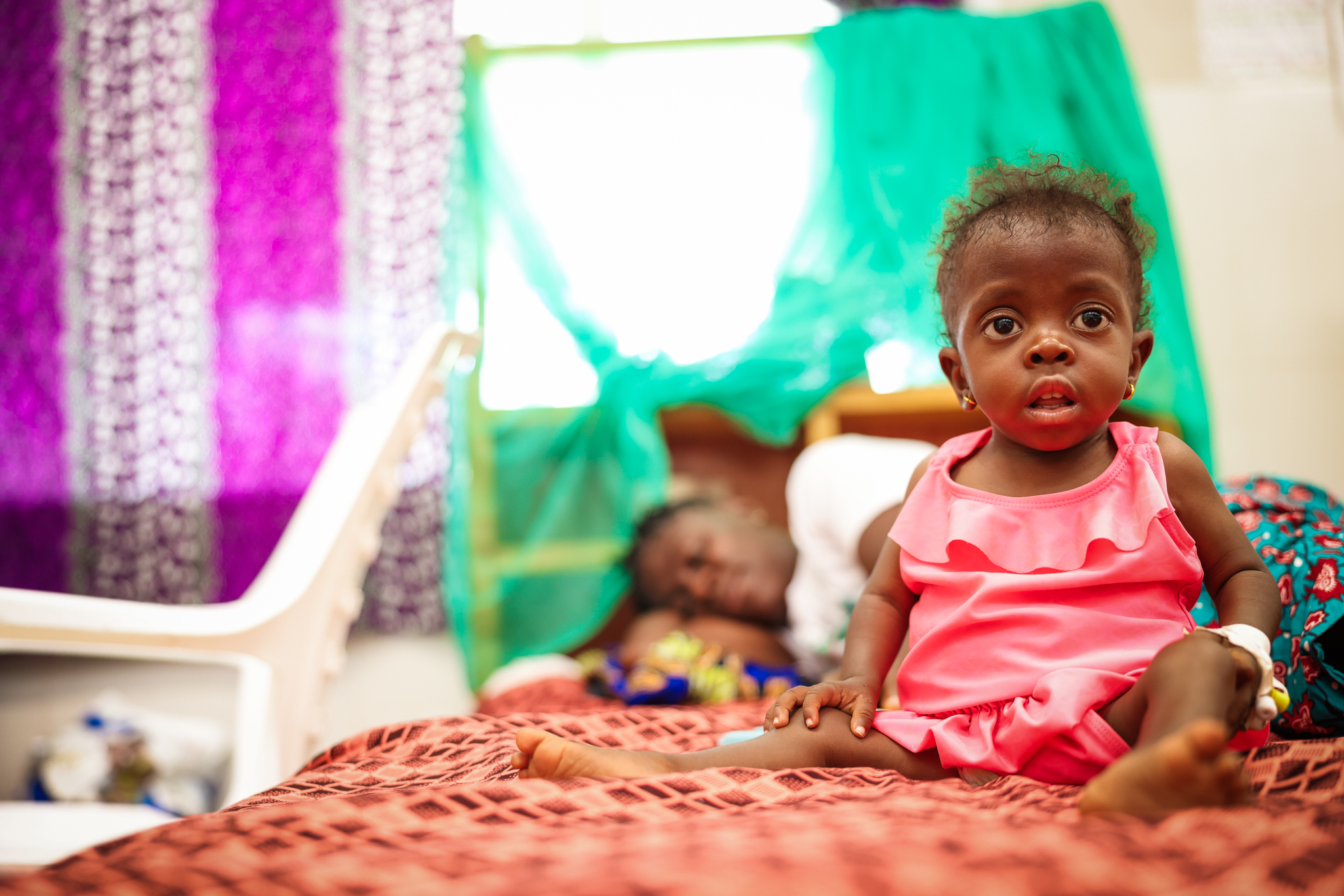 A young girl in a pink dress sits upright and alert on her hospital bed