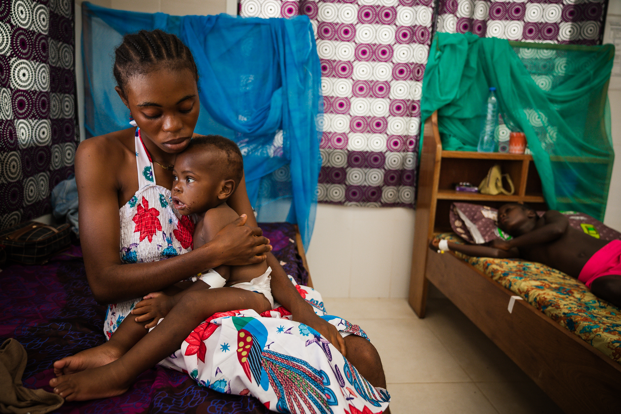 A mother holds her sick child on an inpatient bed, another child resting on a bed behind them